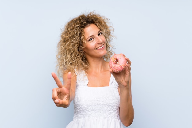 Giovane donna bionda con capelli ricci che tiene una ciambella sopra la parete blu isolata che sorride e che mostra il segno di vittoria