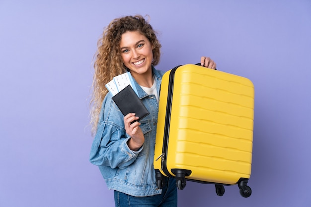 Giovane donna bionda con capelli ricci isolati su viola in vacanza con la valigia e il passaporto