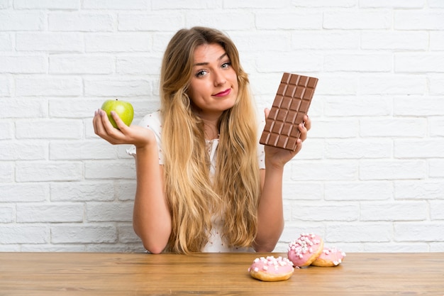 Giovane donna bionda con cioccolato e una mela che ha dubbi