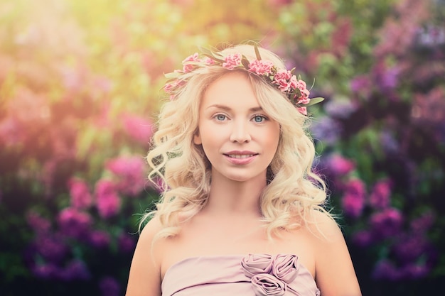 Giovane donna bionda con ghirlanda di fiori e luce solare all'aperto. Bellezza estiva su sfondo floreale