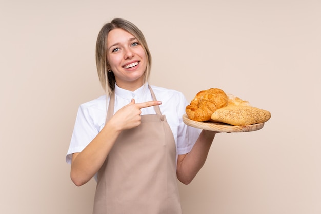 Giovane donna bionda con grembiule. Panettiere femminile che tiene una tabella con parecchi pani e che lo indica