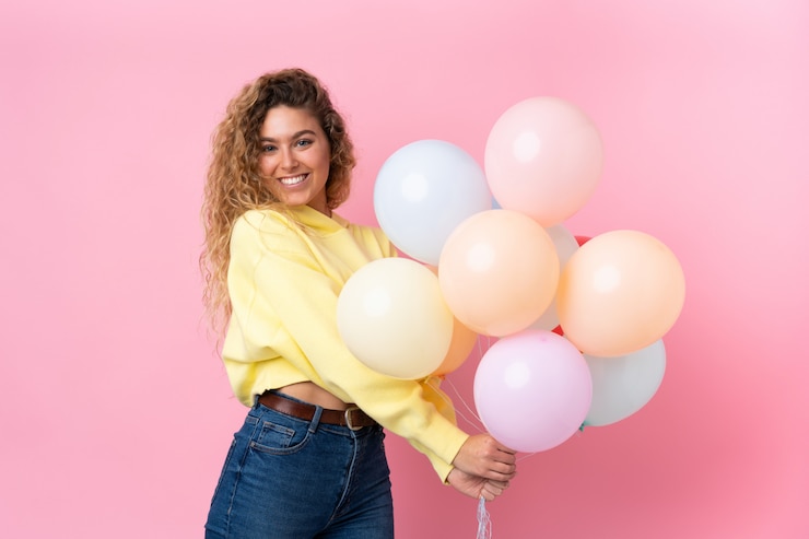 Foto giovane donna bionda con i capelli ricci che cattura molti palloncini sulla parete rosa