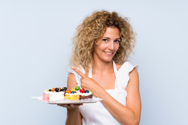 Giovane donna bionda con i capelli ricci in possesso di un sacco di mini torte diverse che punta verso il lato