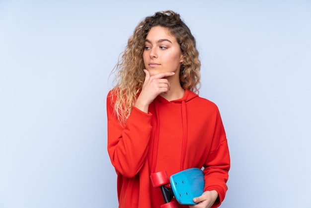 Giovane donna bionda con i capelli ricci sulla parete blu con un pattino e guardando laterale