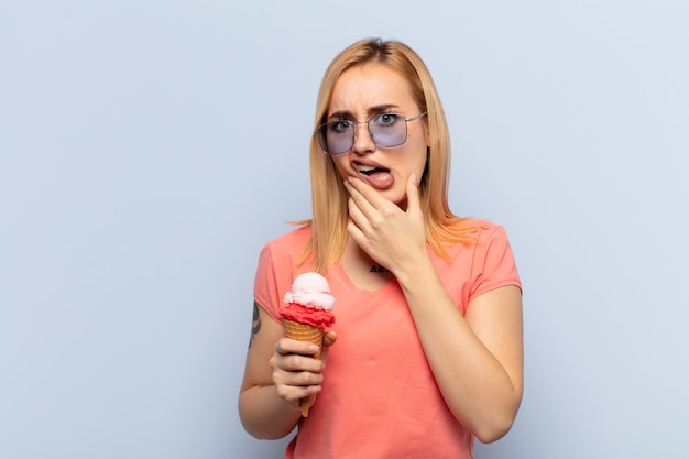 Giovane donna bionda con la bocca e gli occhi spalancati e la mano sul mento, sentendosi spiacevolmente scioccata, dicendo cosa o wow