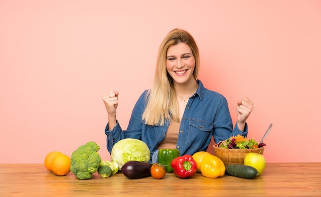 Giovane donna bionda con molte verdure che celebra una vittoria nella posizione del vincitore