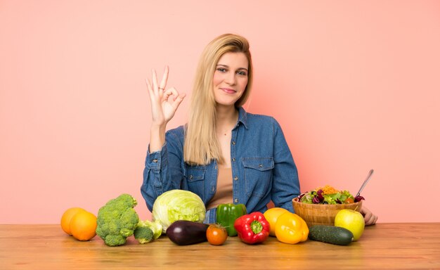 Giovane donna bionda con molte verdure che mostrano segno giusto con le dita