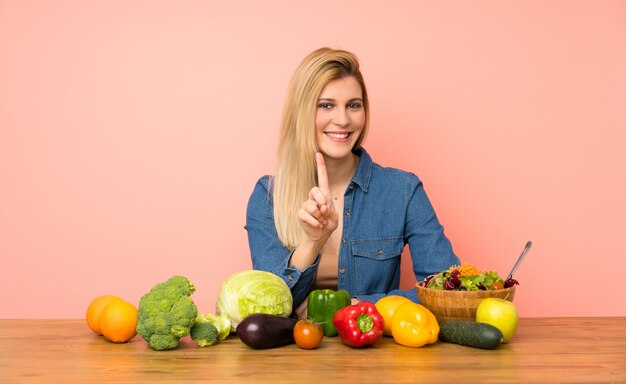 Giovane donna bionda con molte verdure mostrando e alzando un dito