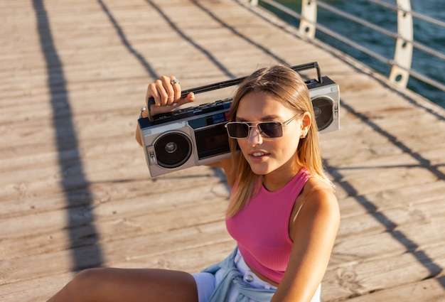 Giovane donna bionda con registratore a nastro retrò sulla spiaggia al luminoso giorno di sole