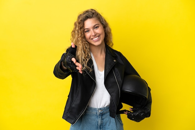 Giovane donna bionda con un casco da motociclista isolato su sfondo giallo che stringe la mano per aver chiuso un buon affare