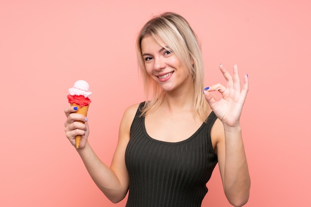 Giovane donna bionda con un gelato di cornetta sopra la parete rosa isolata che mostra segno giusto con le dita