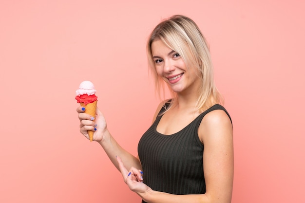 Giovane donna bionda con un gelato di cornetta sopra la parete rosa isolata e indicarla