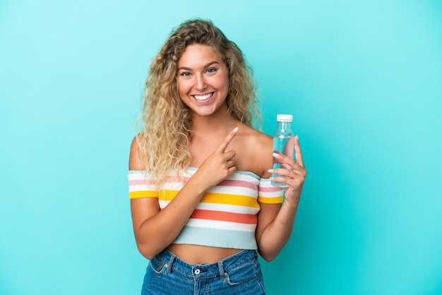 Giovane donna bionda con una bottiglia d'acqua isolata su sfondo blu che indica il lato per presentare un prodotto