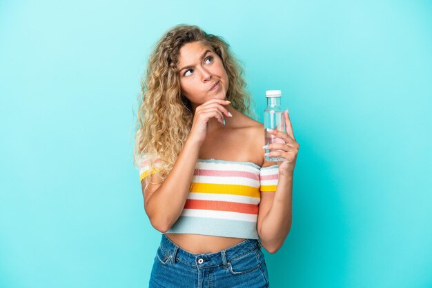 Giovane donna bionda con una bottiglia d'acqua isolata su sfondo blu e alzando lo sguardo