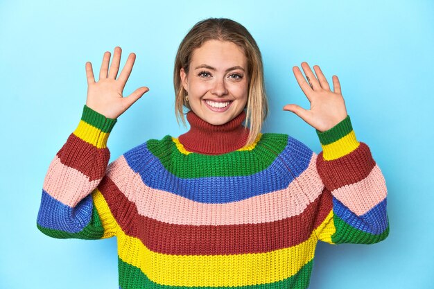 Giovane donna bionda con una felpa multicolore su sfondo blu che mostra il numero dieci con le mani