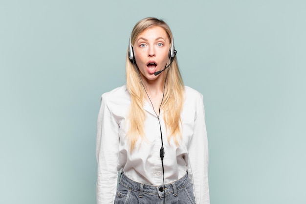 Giovane donna bionda di telemarketing che sembra molto scioccata o sorpresa, fissando con la bocca aperta dicendo wow