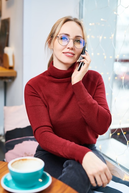 Giovane donna bionda elegante in occhiali e pullover rosso scuro casuale che si siede dalla tavola in caffè e che parla dallo smartphone