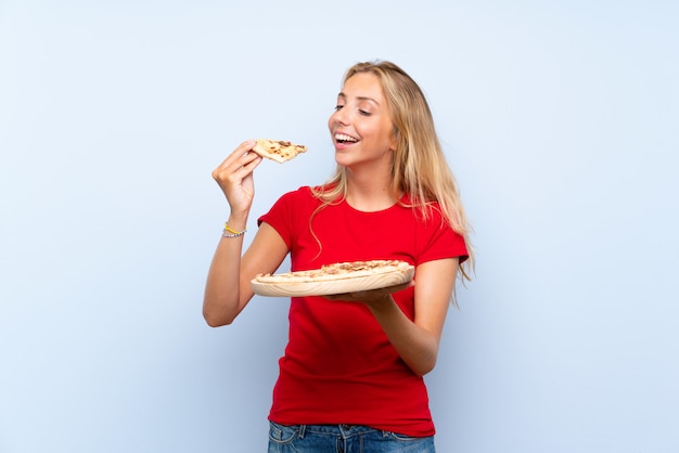 Giovane donna bionda felice che tiene una pizza sopra la parete blu isolata