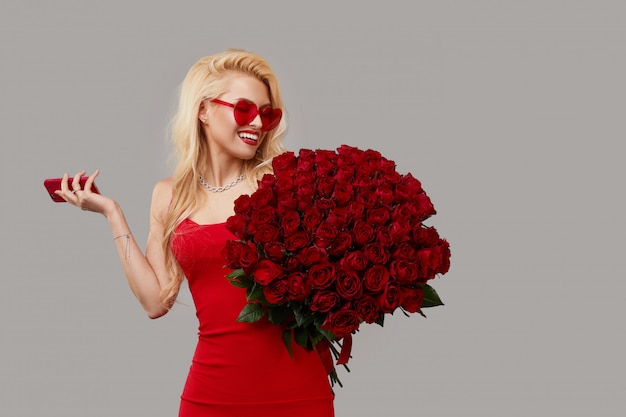 Giovane donna bionda felice in vetri a forma di cuore che tengono un grande mazzo delle rose rosse e del telefono cellulare