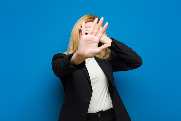 Giovane donna bionda graziosa che copre il viso con la mano e mettendo l'altra mano in alto per fermare la fotocamera, rifiutando foto o immagini contro la parete piatta