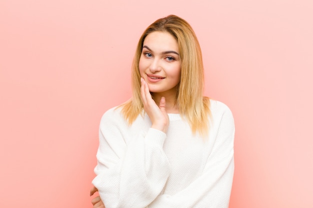 Giovane donna bionda graziosa che sorride, godendo della vita, sentendosi felice, amichevole, soddisfatto e spensierato con la mano sul mento contro la parete piana rosa