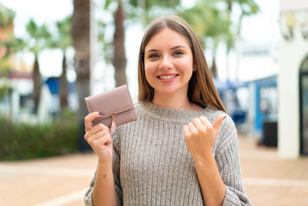 Giovane donna bionda graziosa che tiene un portafoglio all'aperto che indica il lato per presentare un prodotto
