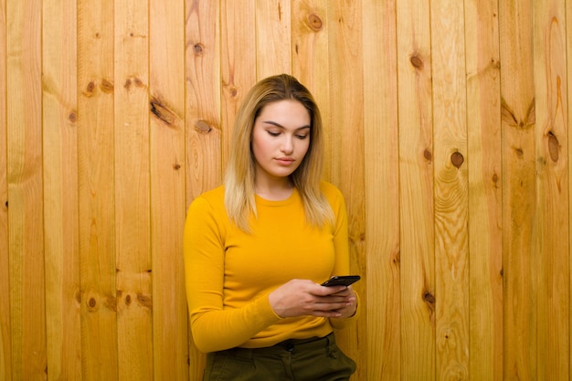 Giovane donna bionda graziosa con un telefono cellulare contro la parete di legno