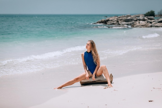 Giovane donna bionda graziosa in un costume da bagno blu in posa su una spiaggia sullo sfondo del mare. Concetto di vacanza estiva