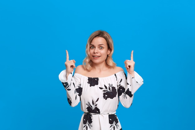 Giovane donna bionda graziosa in un vestito bianco che indica con le dita su mentre stando.