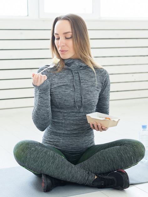 Giovane donna bionda in abbigliamento fitness, con cibo sano vegetariano, seduta sul pavimento