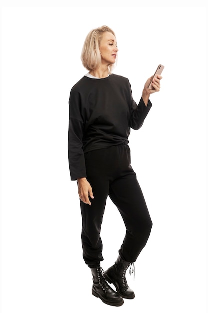 Giovane donna bionda in abito casual nero e stivali ruvidi con un telefono in mano. A tutta altezza. Isolato