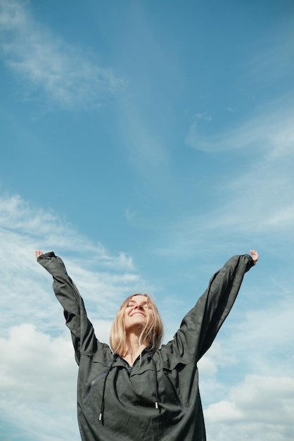 Giovane donna bionda in camicia kaki con le mani in alto davanti al cielo blu all'aperto