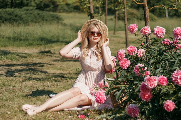 Giovane donna bionda in cappello di paglia vicino all'aiola con le peonie, foto di estate