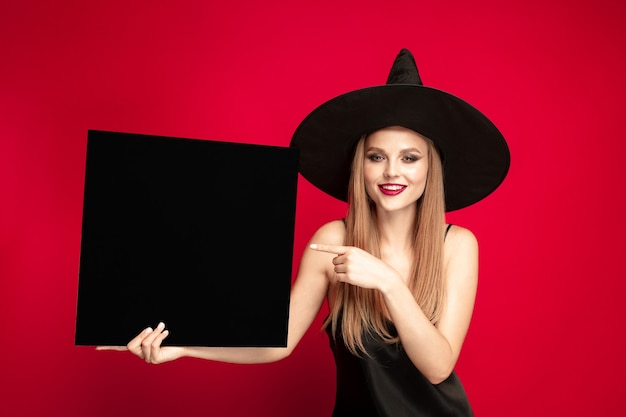 Giovane donna bionda in cappello nero e costume su sfondo rosso. Posa attraente del modello femminile caucasica. Halloween, venerdì nero, cyber lunedì, saldi, concetto autunnale. Copyspace. Contiene targa nera.