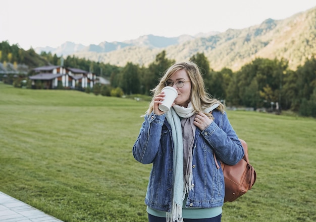 Giovane donna bionda in jeans cappotto con tazza di carta di caffè mattutino al villaggio chalet chalet