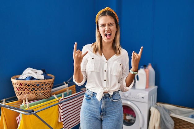 Giovane donna bionda in lavanderia che grida con un'espressione pazza che fa il simbolo rock con le mani in alto. stella della musica. concetto di musica pesante.