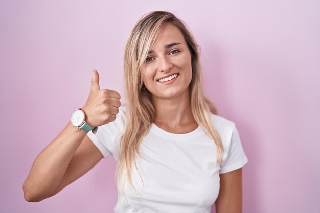 Giovane donna bionda in piedi su sfondo rosa facendo felice pollice in alto gesto con la mano. espressione di approvazione guardando la telecamera che mostra il successo.