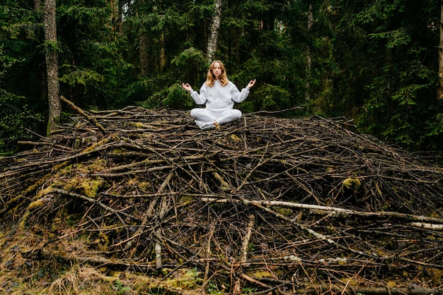 Giovane donna bionda in tuta bianca medita sulla collina del bastone nella foresta