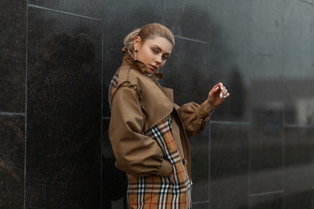 giovane donna bionda in un maglione e un trench beige in posa vicino a un edificio moderno nero