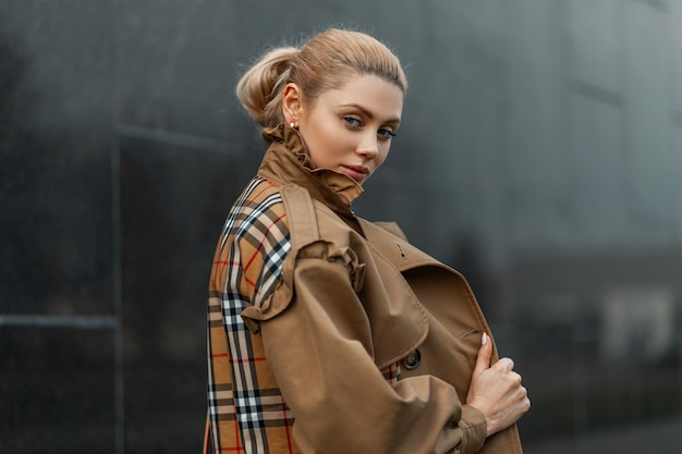 giovane donna bionda in un maglione e un trench beige in posa vicino a un edificio moderno nero