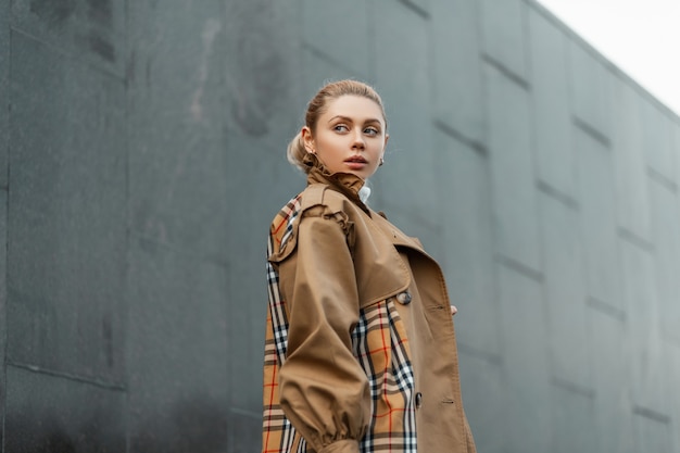 giovane donna bionda in un maglione e un trench beige in posa vicino a un edificio moderno nero