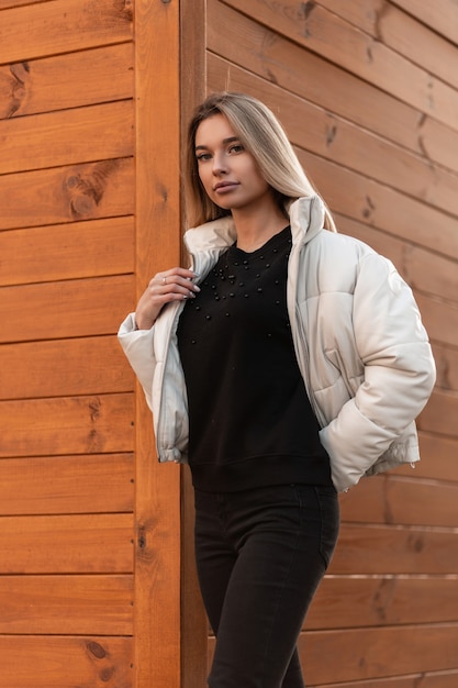 Giovane donna bionda in un maglione nero e una giacca bianca alla moda in posa vicino a un edificio in legno