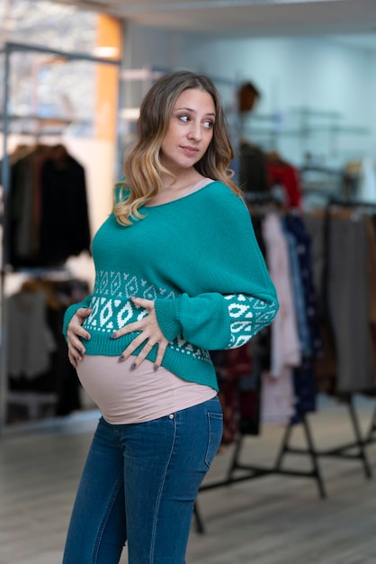 Giovane donna bionda incinta che prova i vestiti in un negozio di abbigliamento Indossa jeans blu e maglione turchese