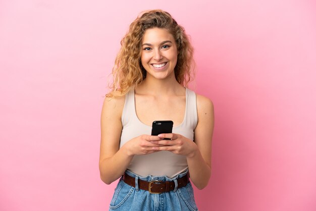 Giovane donna bionda isolata su sfondo rosa guardando la telecamera e sorridendo mentre usa il cellulare