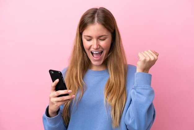 Giovane donna bionda isolata su sfondo rosa utilizzando il telefono cellulare e facendo gesto di vittoria