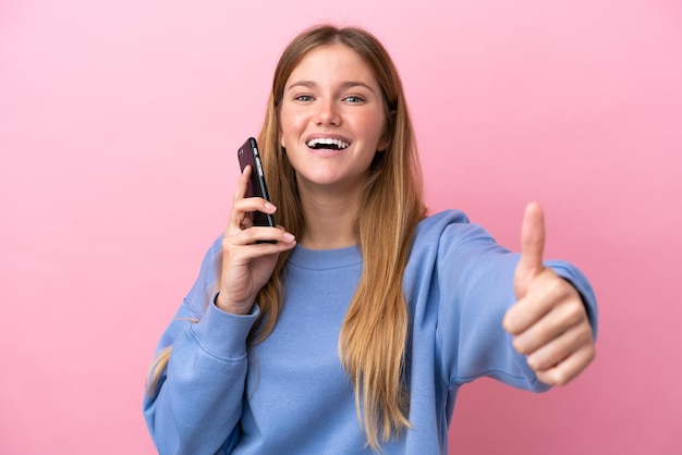 Giovane donna bionda isolata su sfondo rosa utilizzando il telefono cellulare mentre fa i pollici in su