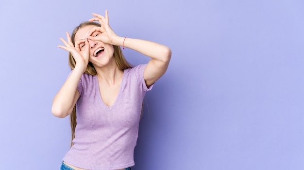 Giovane donna bionda isolata su sfondo viola che mostra bene il segno sugli occhi