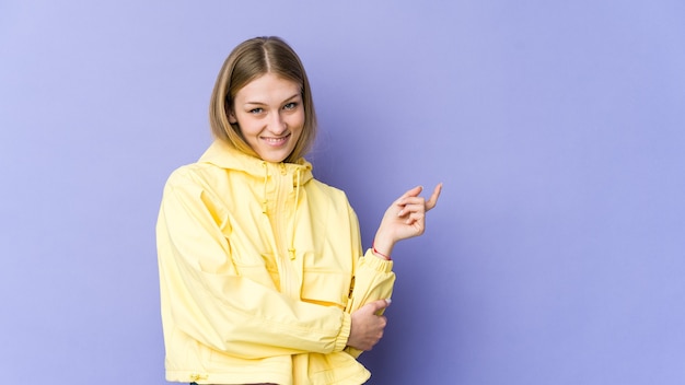 Giovane donna bionda isolata su sfondo viola sorridendo allegramente indicando con l'indice di distanza.