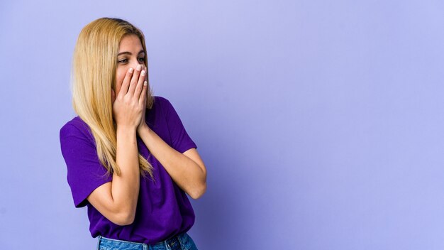 Giovane donna bionda isolata su viola che ride di qualcosa, che copre la bocca con le mani.