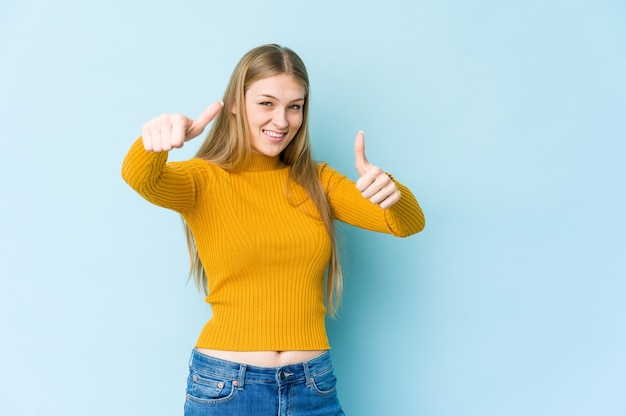 Giovane donna bionda isolata sull'azzurro che alza entrambi i pollici in su, sorridente e sicuro.